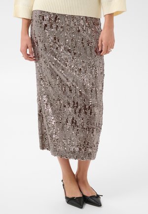 Jupe midi beige à sequins portée avec un pull crème et des ballerines noires pointues, sur une personne debout devant un fond blanc uni.