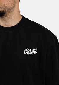 Czarny bawełniany t-shirt z białym stylizowanym logo na górnej lewej części klatki piersiowej. Krótkie rękawy i okrągły dekolt.