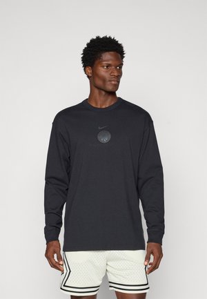 Camisa negra de manga larga de Nike con cuello redondo y parche con logo texturizado en el pecho, combinada con pantalones cortos claros con rayas.