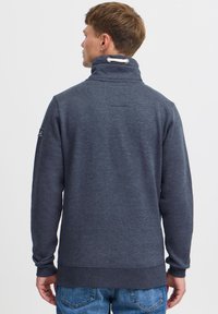 Sudadera de color azul marino con cuello alto, puños y dobladillo acanalados. Presenta un parche con logo en la parte trasera y un acento de cordón en el cuello.