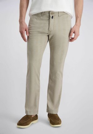 Mann trägt beige legere Hose, weißes Hemd und braune Wildleder-Slipper, steht vor schlichtem hellem Hintergrund.