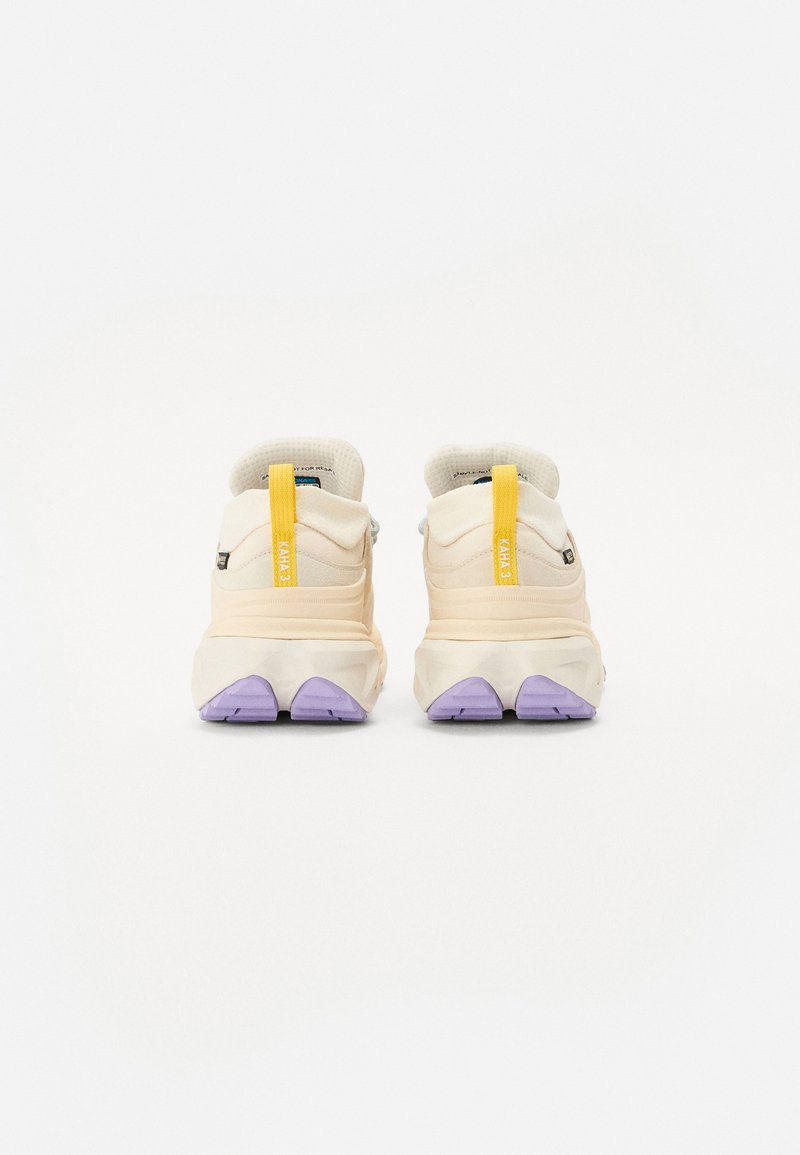Beżowe buty sportowe z teksturowaną cholewką, fioletową podeszwą i żółtym uchwytem na pięcie; zaprojektowane z myślą o komforcie i stabilności.