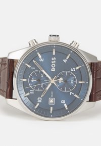 Montre avec un bracelet en cuir brun, un boîtier en acier inoxydable argenté, un cadran bleu, des fonctions chronographe, un affichage de la date à 6 heures et des accents argentés.