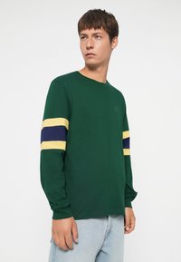 Long-sleeve green t-shirt with yellow and navy stripes on sleeves; round neckline, cotton-like texture, simple design, logo on front.