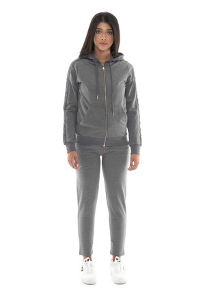Une femme se tient devant un fond blanc, vêtue d’un sweat à capuche gris zippé, d’un pantalon de jogging assorti et de baskets blanches.