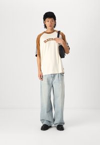 T-shirt avec un bloc de couleurs brun et crème, avec le texte 'RATBAG'. Pantalons en jean bleu clair amples et sac à bandoulière noir. Chaussures noires.