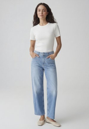 Femme debout, mains dans les poches d’un jean taille haute bleu clair à jambes larges, portant un haut blanc manches courtes et des chaussures plates beiges sur fond gris.