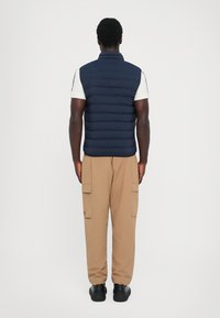 Navy puffer vest with a high collar, stitched detailing, worn over a white shirt, paired with beige cargo pants and black shoes.