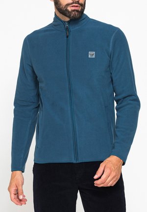 Uomo barbuto che indossa una giacca in pile color verde acqua con zip integrale, con un piccolo logo sul petto sinistro e pantaloni neri.