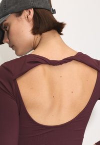 Egy nő bordó felsőt visel, amely nyitott, csavart hátú, és szürke sapkát, amely felfedzi rövid barna haját és csupasz felső hátát.