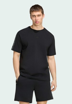 Młody mężczyzna z krótkimi brązowymi włosami, ubrany w czarną koszulkę Adidas z krótkim rękawem oraz pasujące szorty, stoi z jedną ręką w kieszeni.
