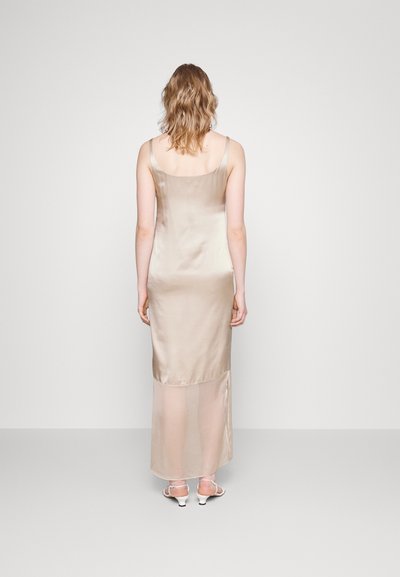 Μακρύ φόρεμα σατέν σε ανοιχτό μπεζ, με λεπτές τιράντες, λεπτομέρεια σχισμής στην πλάτη, ομαλή υφή, συνδυασμένο με λευκά σανδάλια με λουριά.