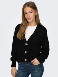 Cardigan preto de malha com decote em V, mangas longas e quatro grandes botões bege. Usado sobre uma camisa branca, combinado com jeans azuis.