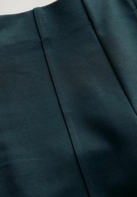 Tmavě modro-zelená tkanina s hladkou texturou, která obsahuje šité švy a svislé linie. Materiál vypadá lehce a jemně.