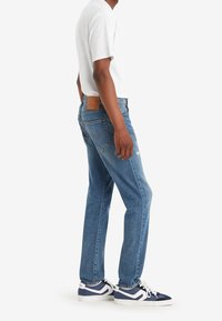 Modré džíny s rovnými nohavicemi, s hnědým koženým štítkem na zadní straně, nošeny s bílým tričkem a námořnickými teniskami.