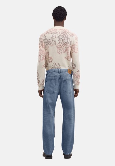 Homme portant un pull léger à motifs floraux et un jean en denim bleu, debout de dos devant un fond uni.