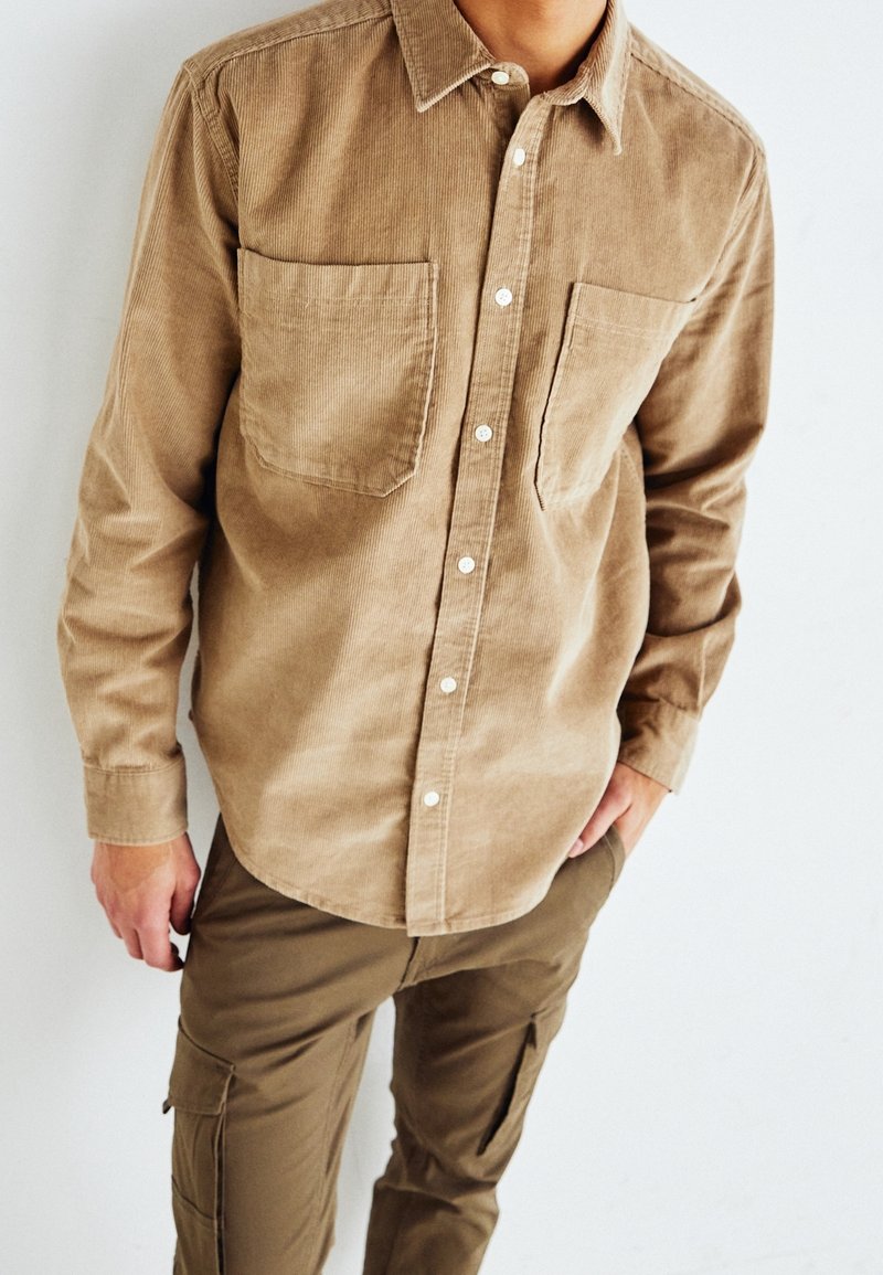 Camicia di velluto a coste marrone con due tasche sul petto, maniche lunghe e bottoni bianchi, abbinata a pantaloni cargo oliva.