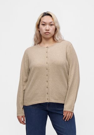 Junge Person mit langen, hellblonden Haaren trägt eine beige, Knopf-Cardigan und dunkelblaue Jeans, steht vor einem schlichten hellen Hintergrund.