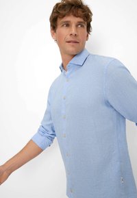 Lichtblauw, langefranje shirt van een gestructureerde stof. Voorzien van een knoopsluiting aan de voorkant met lichtbruine knopen en opgerolde mouwen.