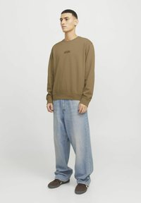 Sudadera marrón de cuello redondo con un pequeño logo impreso en el pecho, combinada con unos jeans de corte holgado en un tono azul claro y zapatillas oscuras marrones.