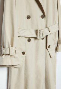 Trench-coat beige avec boutons marron double boutonnage, taille cintrée par ceinture, poches à rabat, et brides ajustables aux poignets des manches longues.