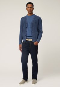 Marineblauwe geribde cardigan met knopen, gedragen over een blauwe crewneck trui. Gecombineerd met donkere spijkerbroeken en lichtgrijze casual schoenen.
