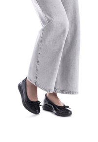 Zapatos de charol negro con punta redonda, que presentan un detalle de lazo en la parte delantera y una parte superior texturizada, combinados con jeans de pierna ancha en gris claro.