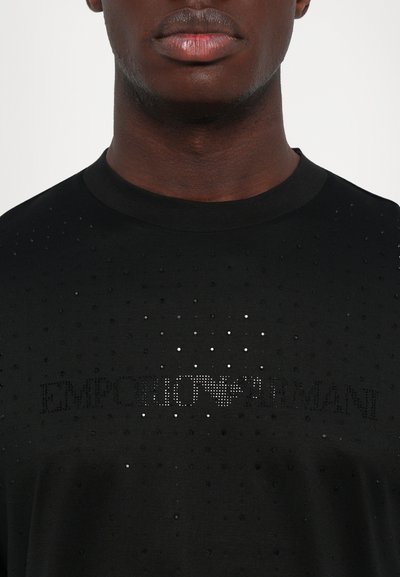 Artimas planas asmens, vilkinčio juodą marškinėlį su mažais blizgančiais kaltiniais, sudarančiais tekstą "EMPORIO ARMANI" per krūtinę.