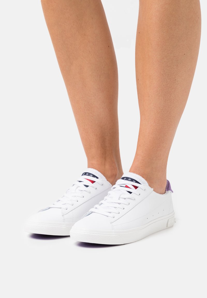 Sneaker bianchi con design a bassa punta, caratterizzati da un materiale in pelle texturizzata, accenti viola sul tallone e dettagli delle stringhe in contrasto.
