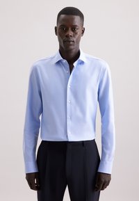 Uomo che indossa una camicia button-down a maniche lunghe blu chiaro e pantaloni neri, in piedi davanti a uno sfondo chiaro e semplice.