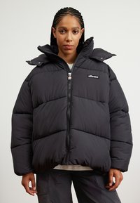 Čierna puffa bunda s vysokým golierom, zipsom vpredu a elastickými manžetami. Obsahuje prešité časti a nášivku s logom na hrudi.