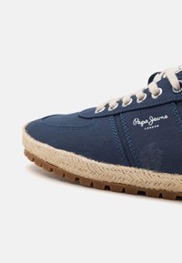 Baskets bleu marine avec semelle tissée beige, lacets blancs et logo "Pepe Jeans London" sur le côté.