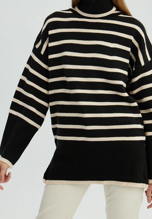 Femme portant un pull à col roulé rayé horizontalement noir et beige avec poignets et ourlet côtelés, associé à un pantalon clair.