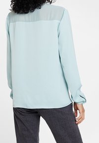 Blouse bleu clair avec un panneau arrière transparent, manches longues et poignets boutonnés. Texture lisse avec une coupe décontractée, associée à un jean foncé.