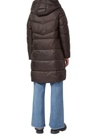 Manteau matelassé marron avec capuche, présentant un matelassage horizontal et une longueur plus longue, associé à un jean bleu clair à jambes larges et des bottes noires.