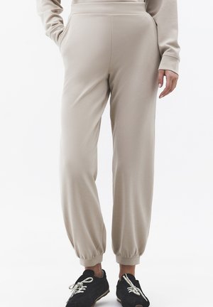 Beige Jogginghosen mit elastischen Bündchen, getragen zu schwarzen Turnschuhen, gezeigt von der Taille bis zu den Füßen.