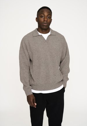 Lys grå, ribbet sweater med V-hals og kort krave, båret med en hvid skjorte under. Iført sorte bukser, stående mod en lys baggrund.