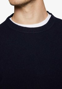 Marineblå strikketrøje med rund halsudskæring og ribbet krave. Lagt over en hvid T-shirt. Materialet virker blødt med en glat tekstur.