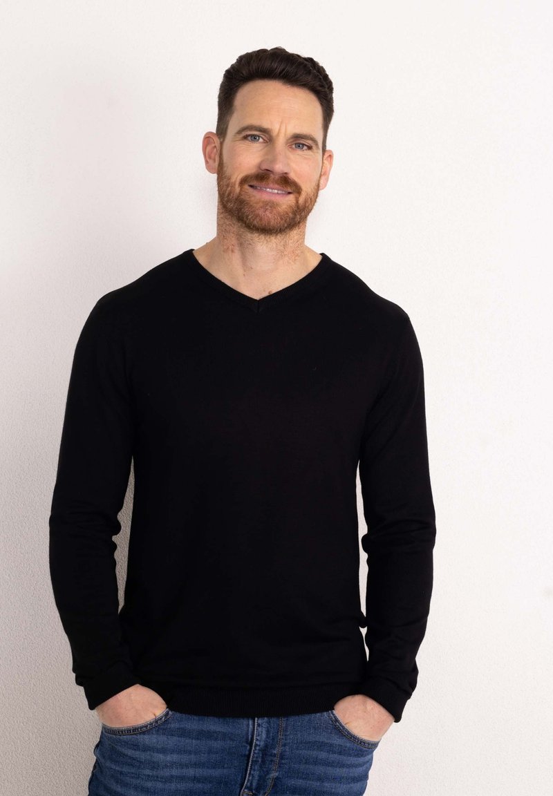 Maglione nero a maniche lunghe con scollo a V, materiale in maglia morbido, polsini a costine, abbinato a jeans azzurri chiari, indossato in modo casual con le mani nelle tasche.