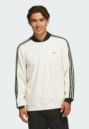 Young man smiling wearing a white Adidas sweatshirt with black stripes on sleeves and black collar and cuffs.