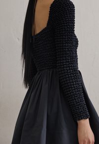 Svart klänning med smockad bodice, långa ärmar och en flödande kjol. Tyget har en slät textur med rynkade detaljer.