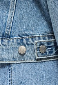 Κοντινό πλάνο σε μπλε denim μπουφάν που δείχνει δύο μεταλλικά κουμπιά και λεπτομέρειες ραφής στον ιμάντα ρύθμισης μέσης.