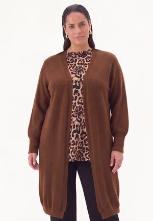 Femme portant un long cardigan marron par-dessus un haut à imprimé léopard et un pantalon noir, debout devant un fond clair uni.