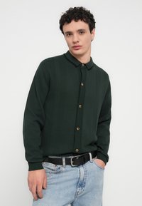 Πράσινη σκούρη πλεκτή ζακέτα με κάθετες ρίγες, μαύρο γιακά και ξύλινα κουμπιά, συνδυασμένη με ανοιχτόχρωμο μπλε τζιν και μαύρη ζώνη.