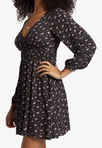 Žena v černých květinových šatech s dlouhými rukávy, výstřihem do V a nařaseným pasem, pózuje s jednou rukou na bocích.