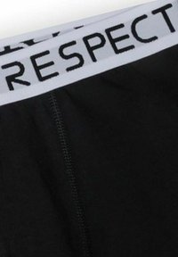 Fekete anyag fehér derékrésszel, amelyen a "TISZTELET" szó vastag fekete betűkkel olvasható, és látható varrás található alatta.