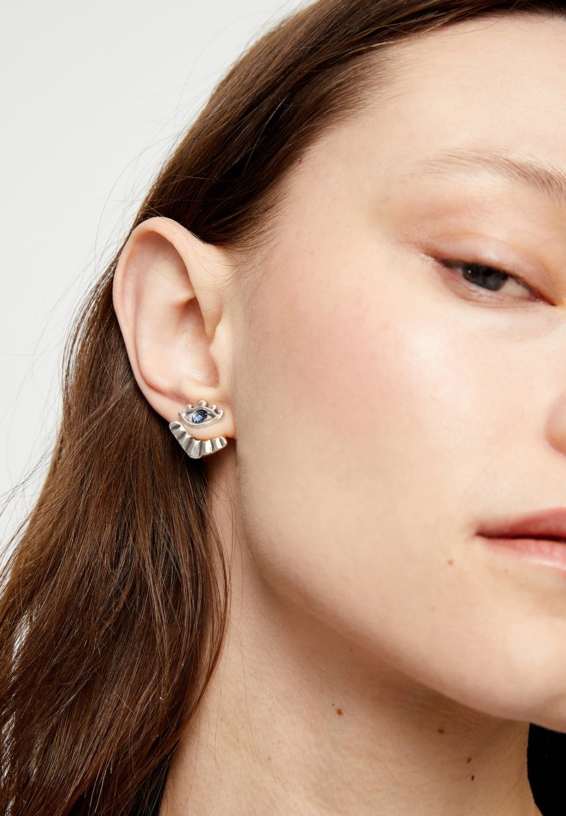 Boucles d'oreilles en argent avec un motif d'œil orné d'une pierre précieuse bleue. Un accent texturé ondulé complète la forme, mettant en valeur un savoir-faire unique.