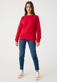 Sudadera roja de manga larga, cuello redondo y dobladillo acanalado, combinada con jeans ajustados azules y zapatillas blancas. Tejido suave y diseño casual.