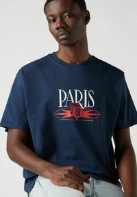 Camiseta de algodón azul marino con un estampado gráfico que presenta "PARÍS" y un diseño de un monumento en rojo, con detalles redondeados. Manga corta, ajuste relajado.