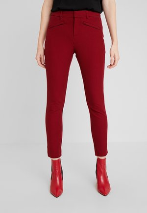 Kvinde iført tætsiddende røde ternede bukser kombineret med røde spidse ankelstøvler, stående mod en ensartet lys baggrund.